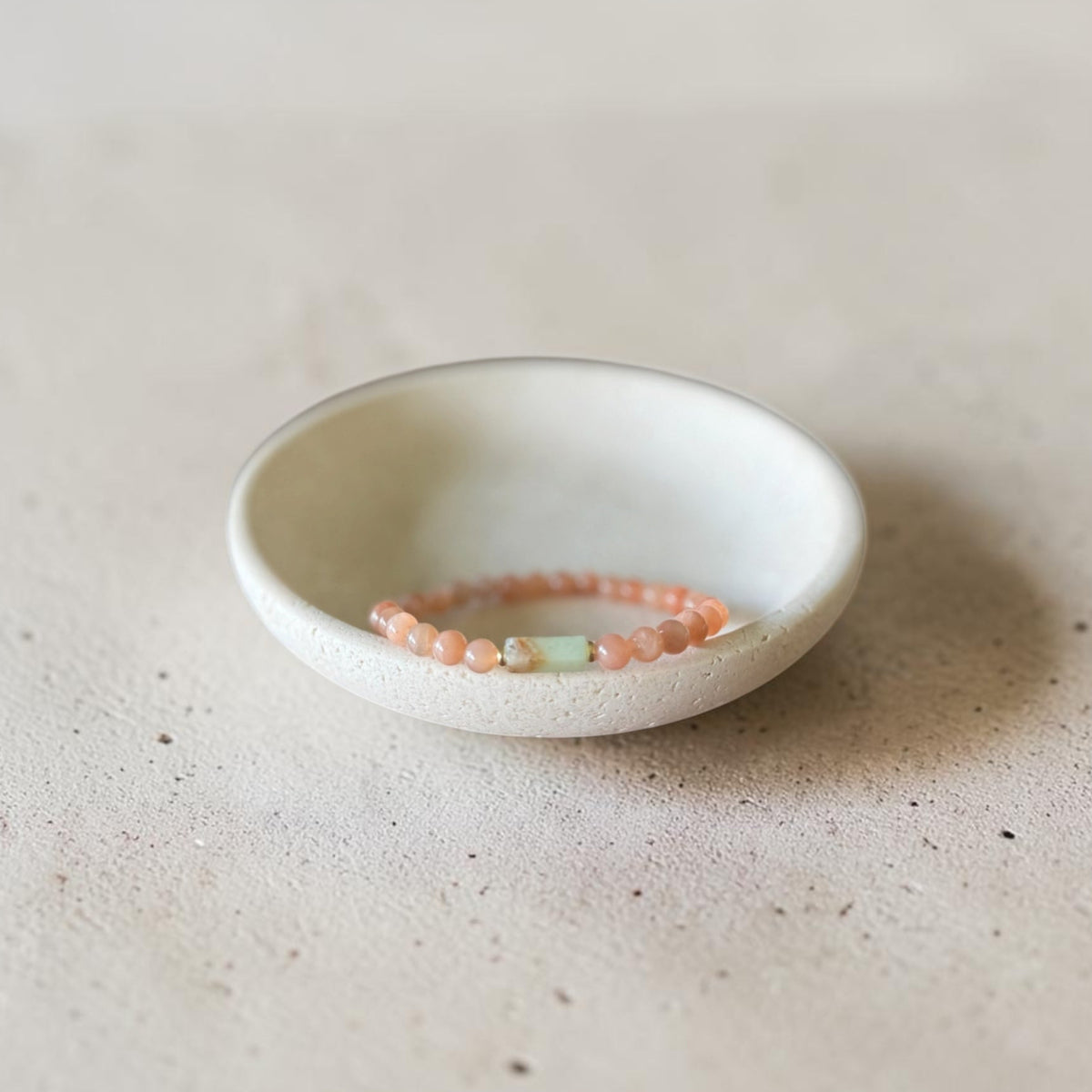 A delicate beaded bracelet in soft peach-pink and pale green tones rests inside a small, shallow white ceramic dish on a textured beige surface.