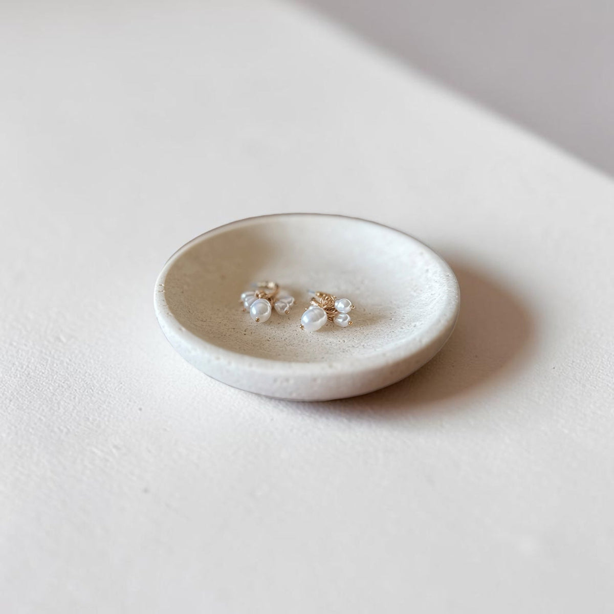 A pair of small gold stud earrings with clear crystal-like floral clusters, displayed in a shallow round white dish on a soft beige surface.