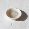 A small round ceramic dish or saucer with a matte, speckled off-white finish, photographed on a textured white surface with a soft shadow cast to the right.  Inside sits a wire bracelet in cream opal color.