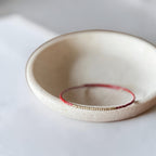 A delicate beaded bracelet with red and gold beads rests inside a small, textured beige ceramic dish on a soft white surface.