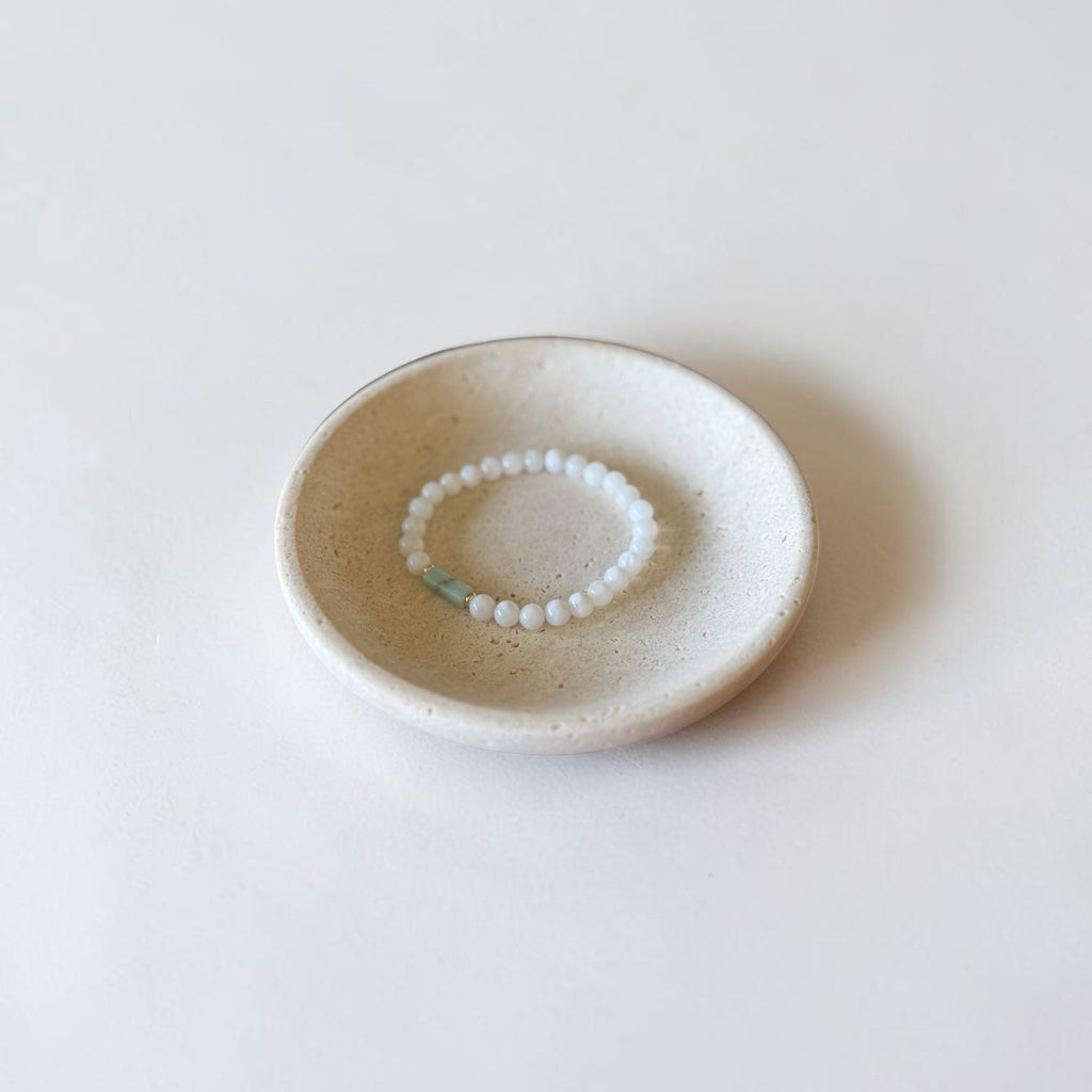 A small white beaded bracelet with a tiny light blue-green bead, displayed inside a round, speckled beige ceramic dish on a clean white background.