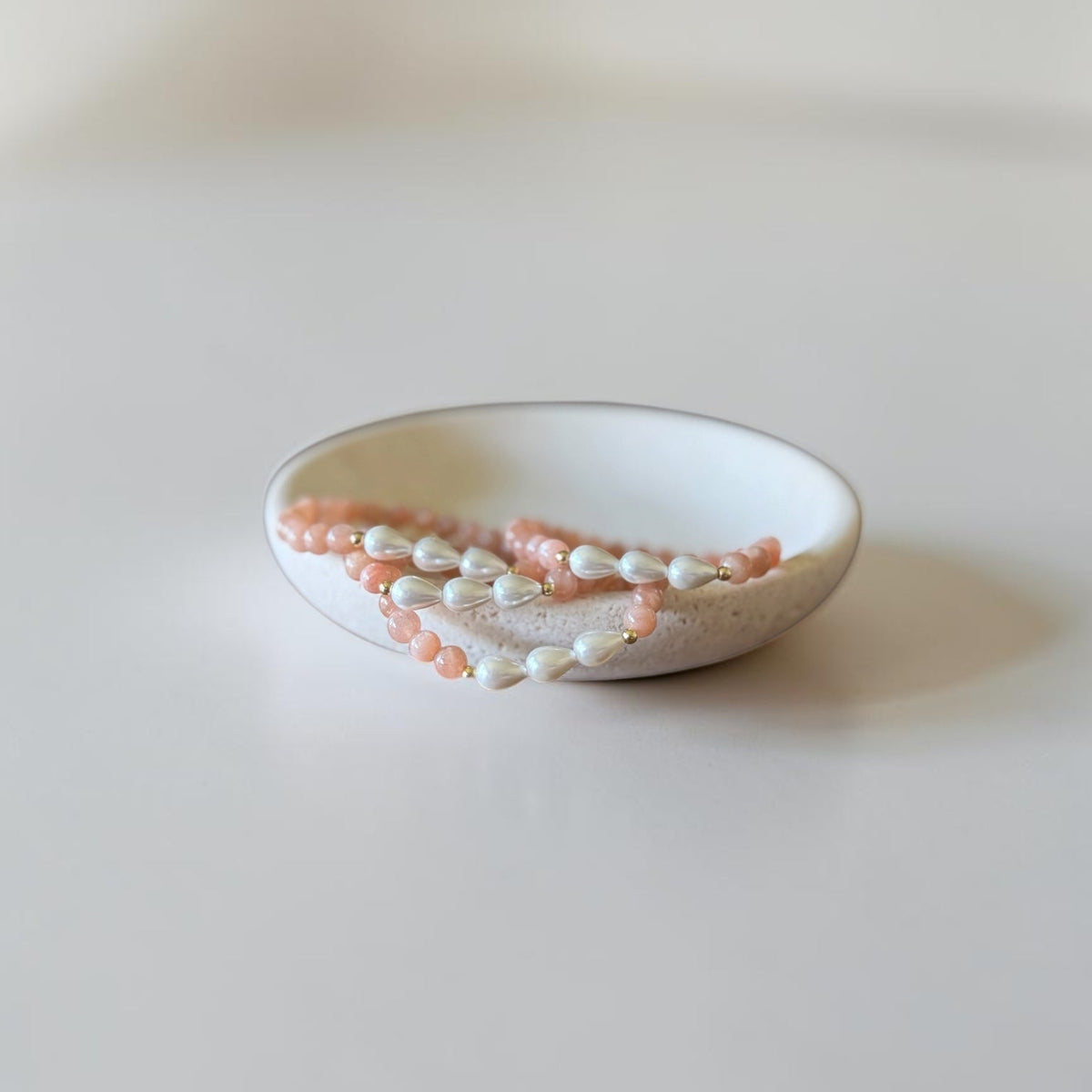 A small white ceramic dish holding several strands of pale pink and white beaded jewelry on a soft neutral background.