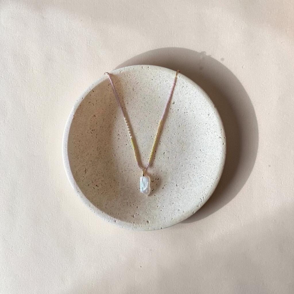 A delicate necklace with a small white irregular pendant and pale pink and gold-toned chain, displayed on a round textured ceramic dish against a warm beige background.