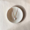 A delicate necklace with a small white irregular pendant and pale pink and gold-toned chain, displayed on a round textured ceramic dish against a warm beige background.
