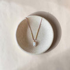 A delicate necklace with a small white irregular pendant and pale pink and gold-toned chain, displayed on a round textured ceramic dish against a warm beige background.