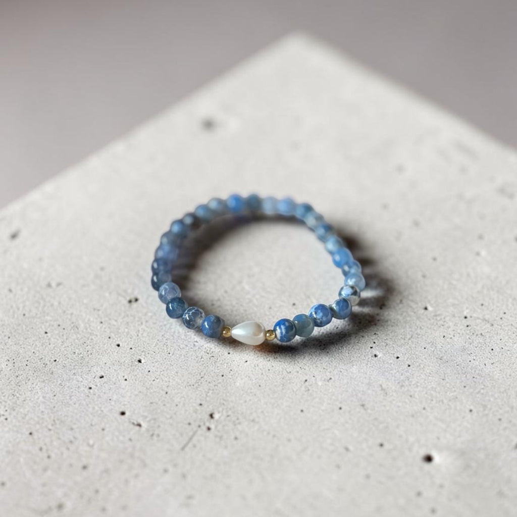 A beaded stretch bracelet featuring light blue faceted beads and a central white teardrop bead with a small gold accent, displayed on a textured light gray concrete surface.