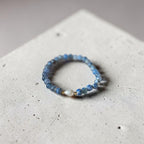 A beaded stretch bracelet featuring light blue faceted beads and a central white teardrop bead with a small gold accent, displayed on a textured light gray concrete surface.