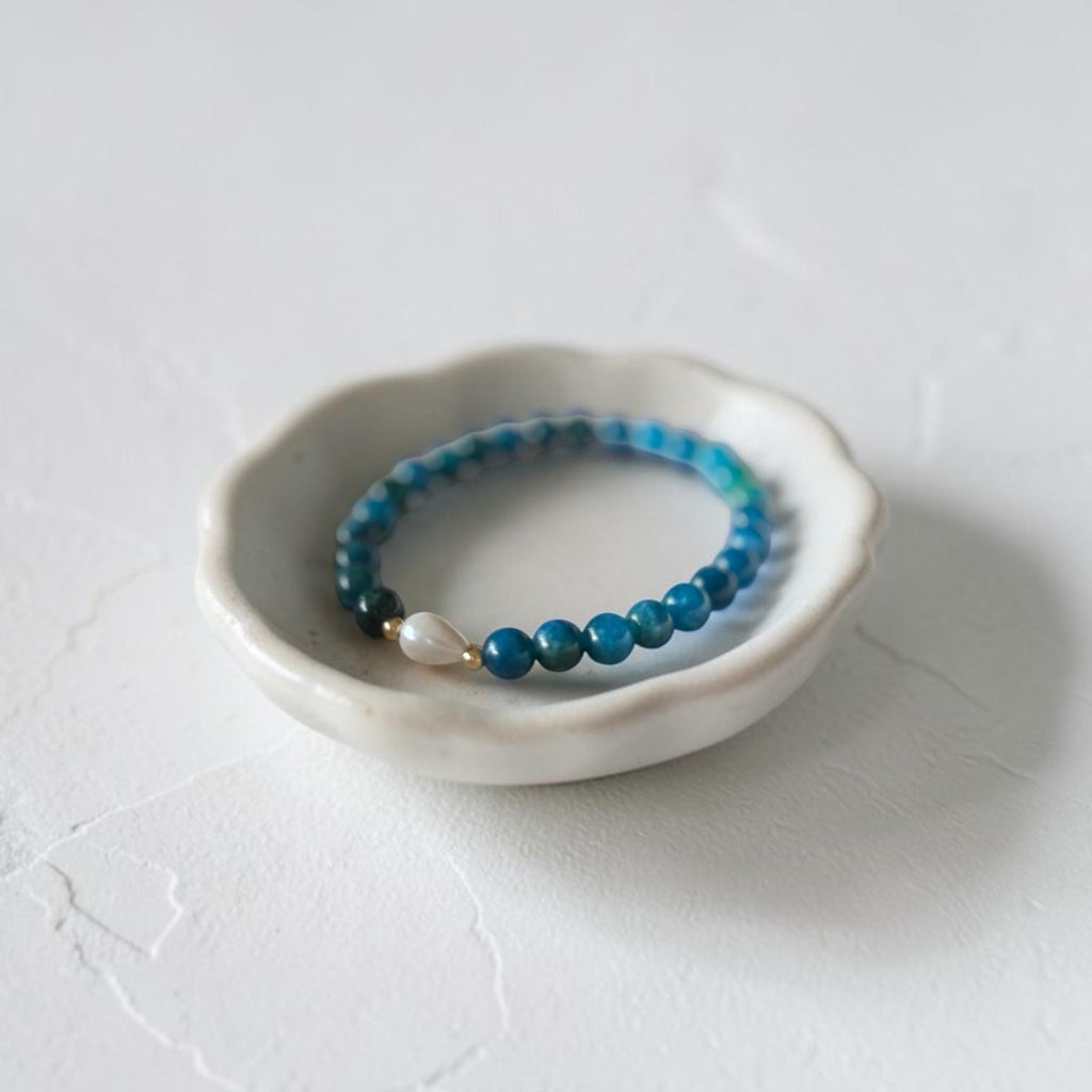 A beaded stretch bracelet in shades of blue and teal with a small white pearl accent, displayed in a small white ceramic dish on a textured white surface.