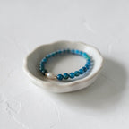 A beaded stretch bracelet in shades of blue and teal with a small white pearl accent, displayed in a small white ceramic dish on a textured white surface.