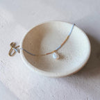 A delicate beaded necklace with a white pearl pendant resting in a small speckled ceramic dish on a textured light gray surface.