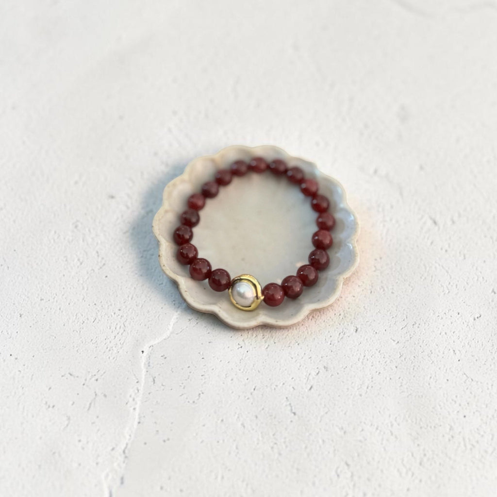 A beaded bracelet with dark red faceted beads and a central gold ring holding a small white pearl, displayed on a small scalloped ceramic dish against a textured off-white background.