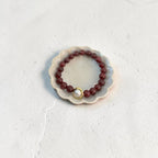 A beaded bracelet with dark red faceted beads and a central gold ring holding a small white pearl, displayed on a small scalloped ceramic dish against a textured off-white background.