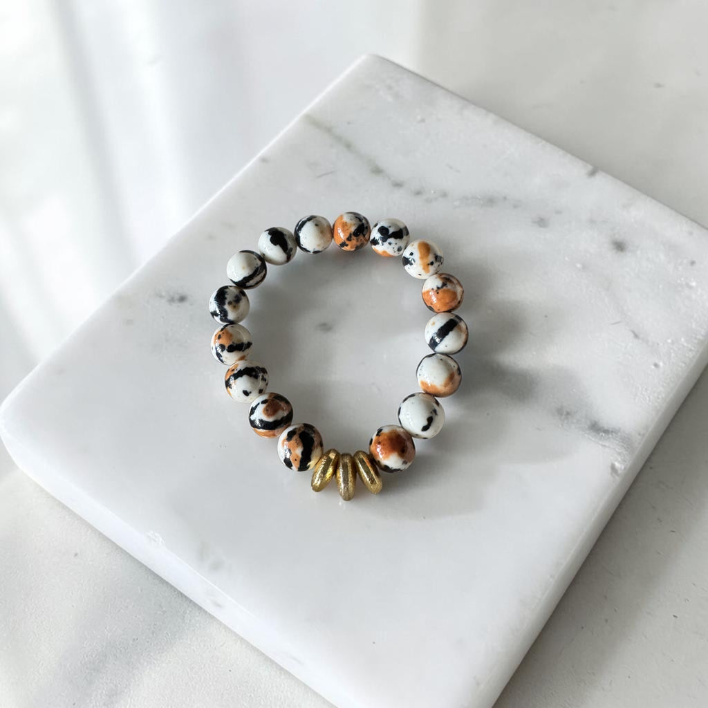 A beaded stretch bracelet with white, black, and orange marbled beads and a small gold-toned accent bead, displayed on a white marble square against a light marble surface.