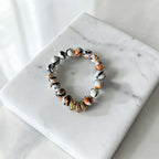 A beaded stretch bracelet with white, black, and orange marbled beads and a small gold-toned accent bead, displayed on a white marble square against a light marble surface.
