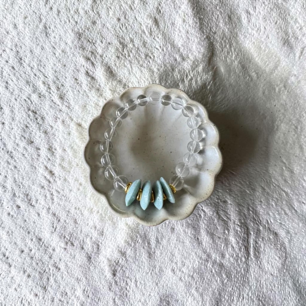A clear beaded bracelet with three light blue teardrop-shaped beads and small gold accents, displayed in a small scalloped white dish on a textured white fabric background.
