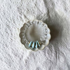 A clear beaded bracelet with three light blue teardrop-shaped beads and small gold accents, displayed in a small scalloped white dish on a textured white fabric background.