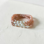 A beaded wrap-style bracelets featuring peach-pink round beads and a central row of white baroque pearls separated by tiny gold spacers, resting on a textured off-white surface.
