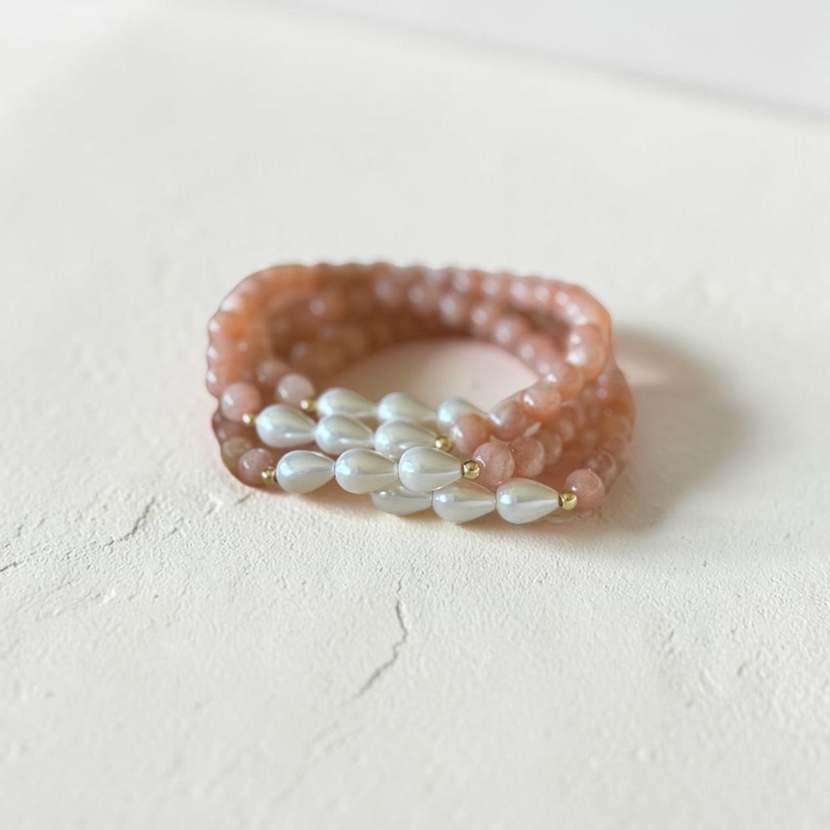 A beaded wrap-style bracelets featuring peach-pink round beads and a central row of white baroque pearls separated by tiny gold spacers, resting on a textured off-white surface.
