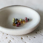 A small round off-white ceramic dish with a speckled texture, holding a few small colorful glass beads on a matching textured surface.