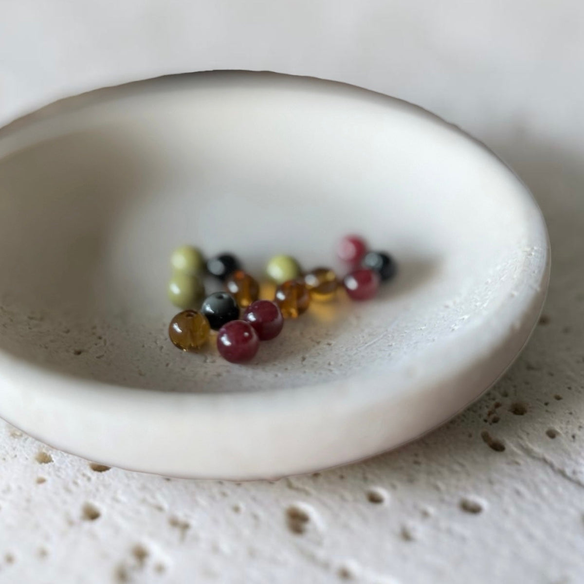 A small round off-white ceramic dish with a speckled texture, holding a few small colorful glass beads on a matching textured surface.