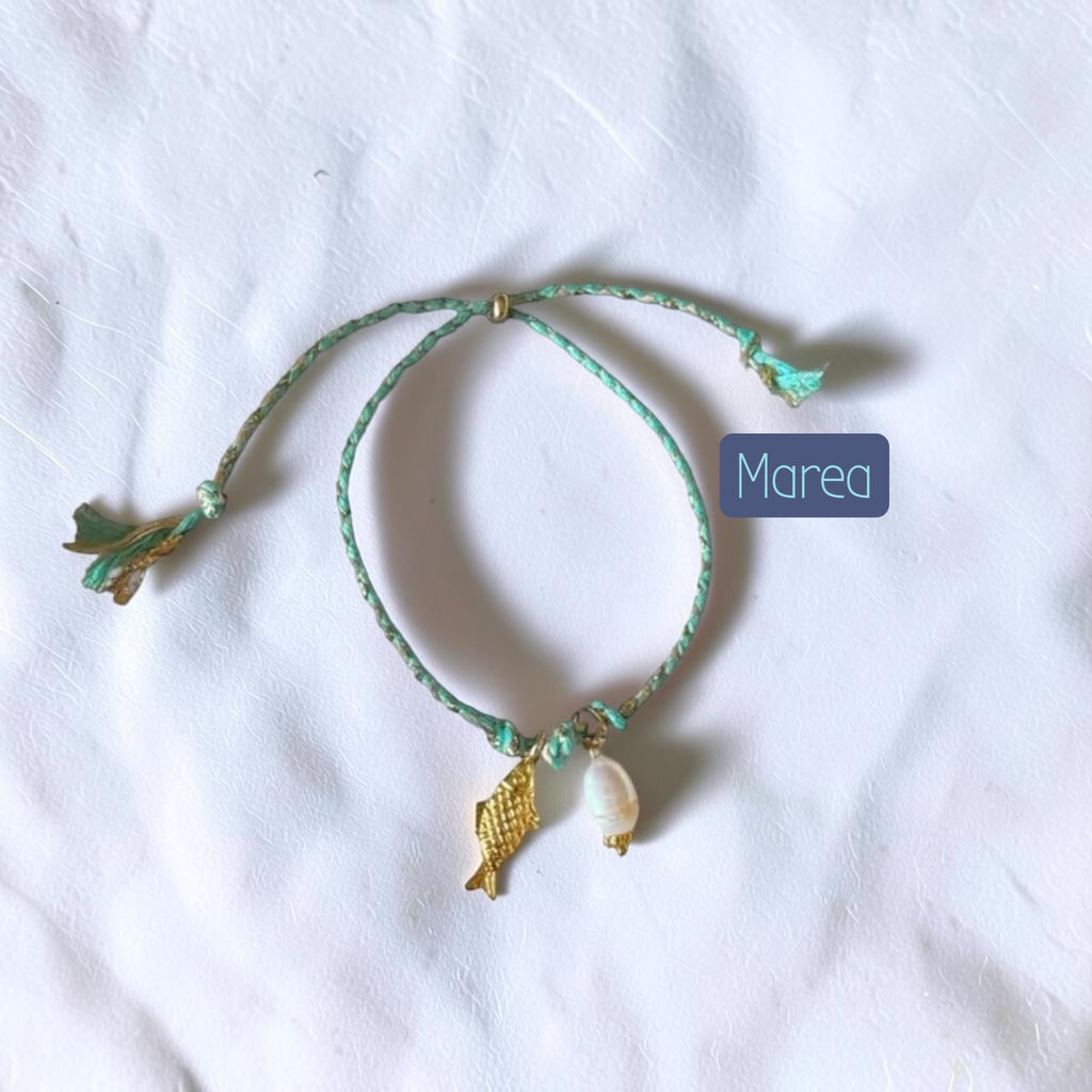 A mint green braided cord bracelet with a small gold fish charm, a white shell charm, and a small gold bead, displayed on a white fabric background with a 'Marea' label overlay.