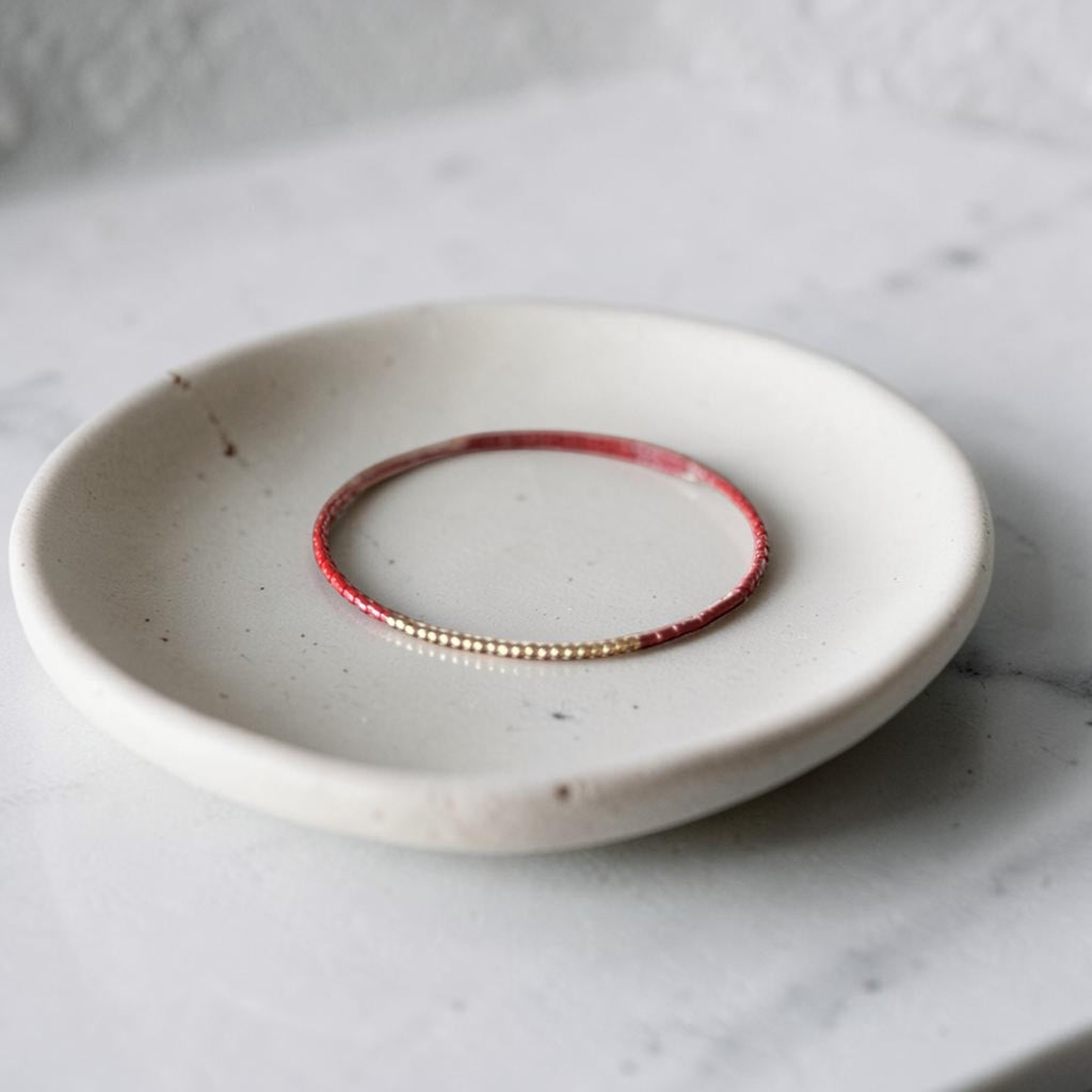 A delicate beaded bracelet with alternating red and gold beads, displayed on a small speckled ceramic dish over a light marble surface.