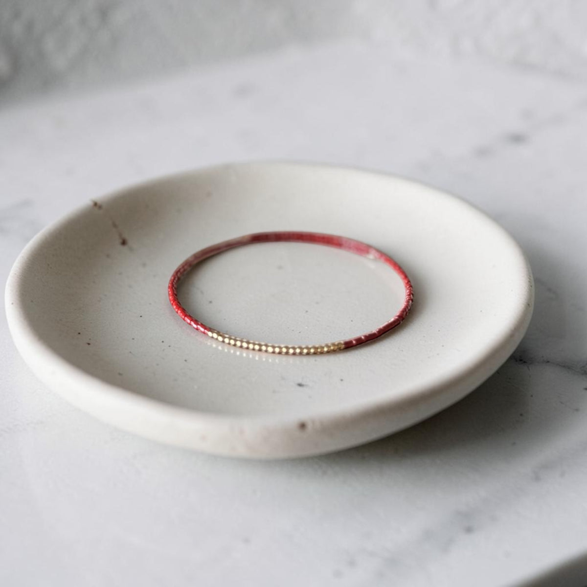 A delicate beaded bracelet with alternating red and gold beads, displayed on a small speckled ceramic dish over a light marble surface.