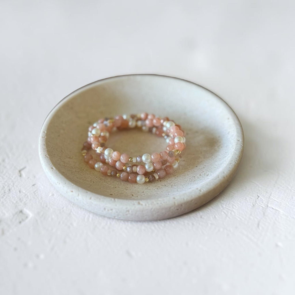 A stack of three beaded stretch bracelets in pastel pink, peach, and white, displayed on a small round ceramic dish against a textured off-white background.