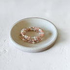 A stack of three beaded stretch bracelets in pastel pink, peach, and white, displayed on a small round ceramic dish against a textured off-white background.