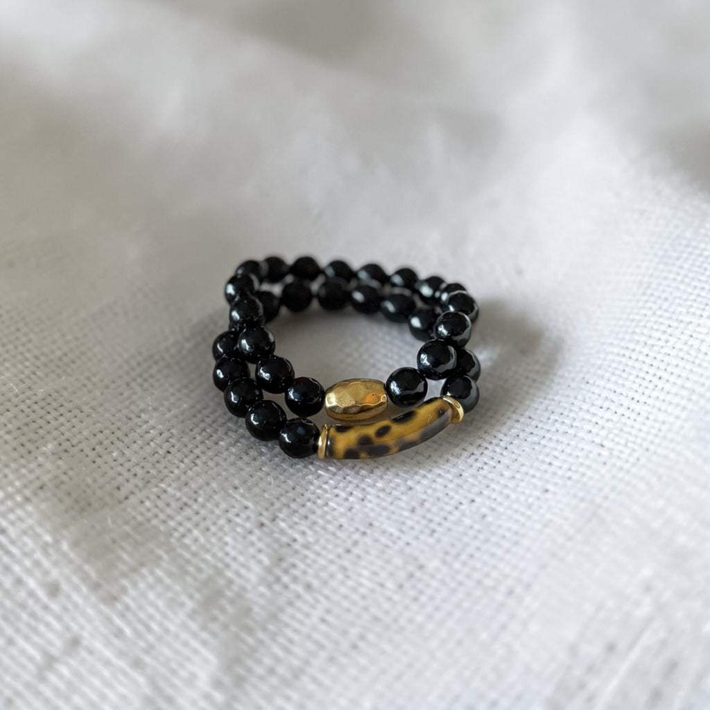 Two beaded bracelets featuring glossy black beads and a central gold-toned spacer bead with a brown tiger-eye pattern, resting on a textured white fabric background.
