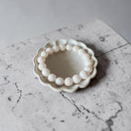 A beaded bracelet made of 10mm round, off-white beads arranged in a circle inside a small, scalloped ceramic dish, set on a gray marble surface.