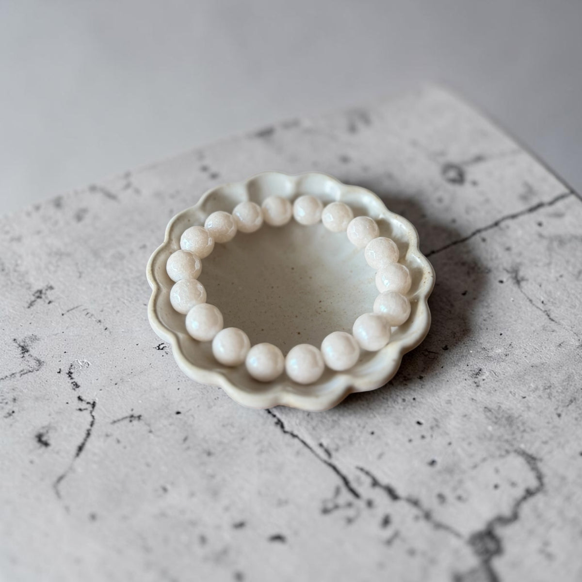 A beaded bracelet made of 10mm round, off-white beads arranged in a circle inside a small, scalloped ceramic dish, set on a gray marble surface.