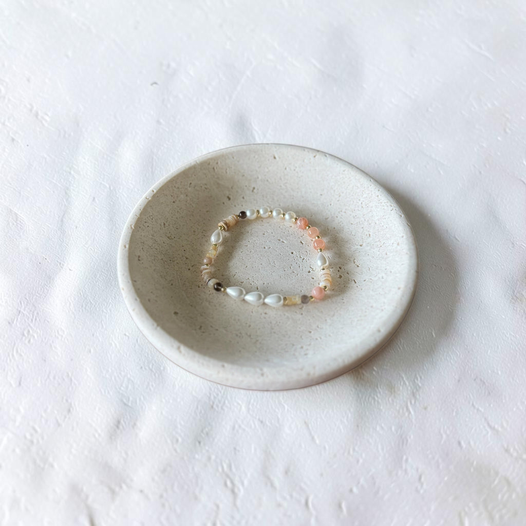 A delicate beaded bracelet with white, peach, and dark brown beads, displayed inside a small speckled ceramic dish on a textured white fabric background.