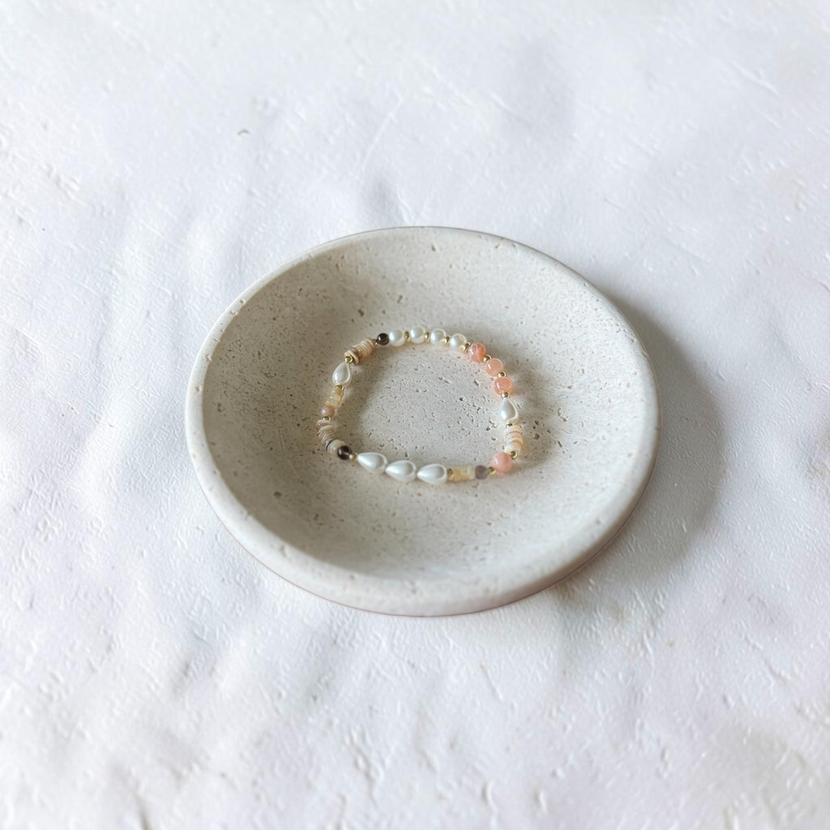 A delicate beaded bracelet with white, peach, and dark brown beads, displayed inside a small speckled ceramic dish on a textured white fabric background.