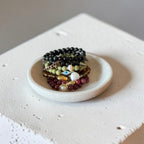 A stack of beaded bracelets in black, green, red, and gold tones displayed on a small round white dish, set on a textured white block against a neutral gray background.