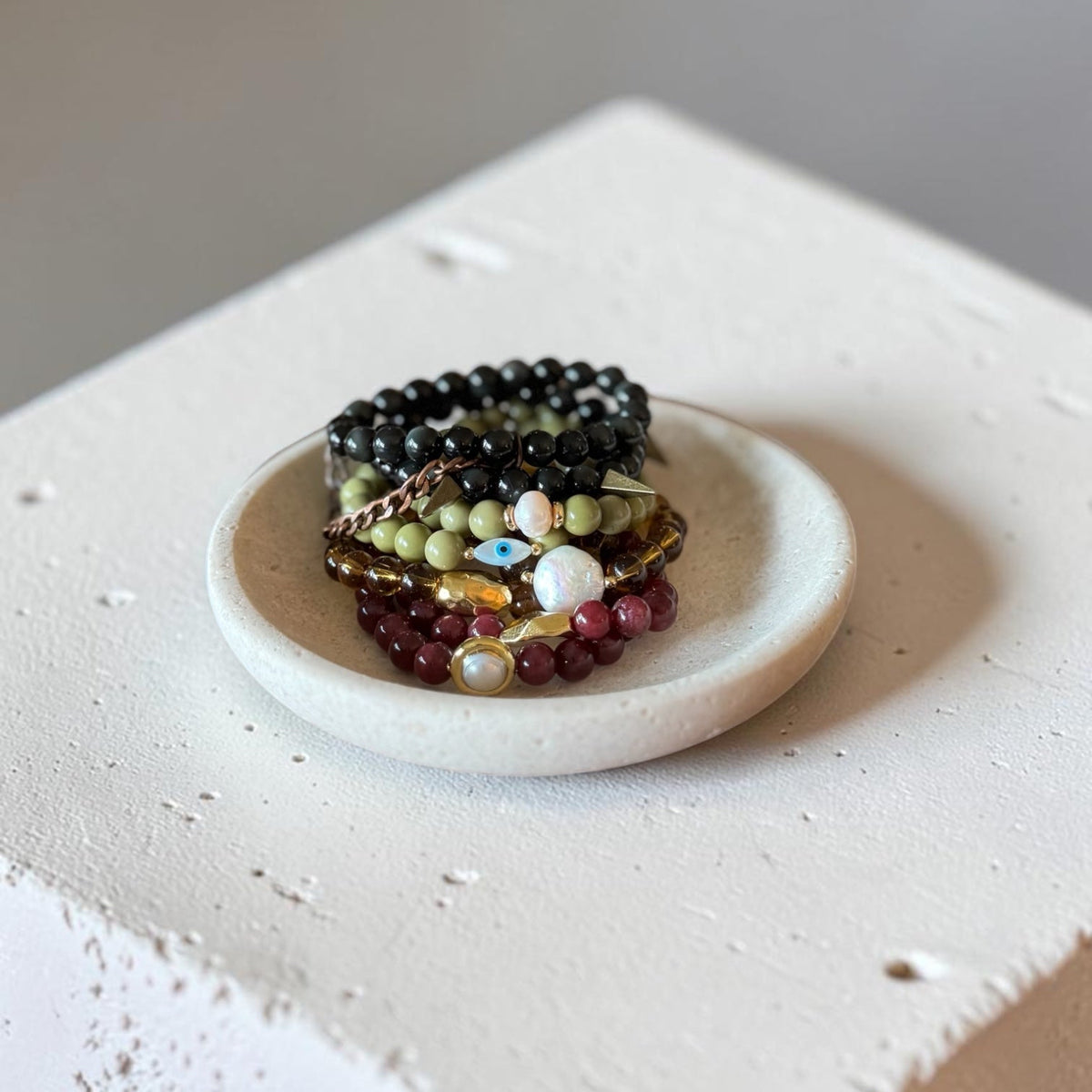 A stack of beaded bracelets in black, green, red, and gold tones displayed on a small round white dish, set on a textured white block against a neutral gray background.