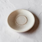 A small round ceramic dish with a textured, speckled off-white glaze, photographed from a top-down angle on a light, textured surface.