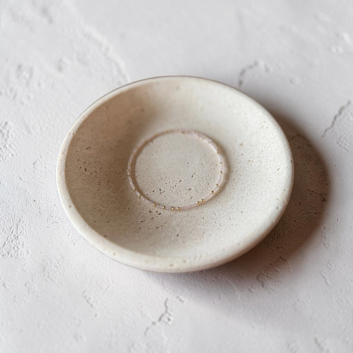 A small round ceramic dish with a textured, speckled off-white glaze, photographed from a top-down angle on a light, textured surface.