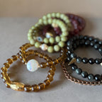 A group of beaded bracelets arranged on a neutral surface. The set includes translucent amber-yellow beads with a white iridescent bead, light olive-green beads, dark charcoal gray beads, and a rose-gold chain bracelet.