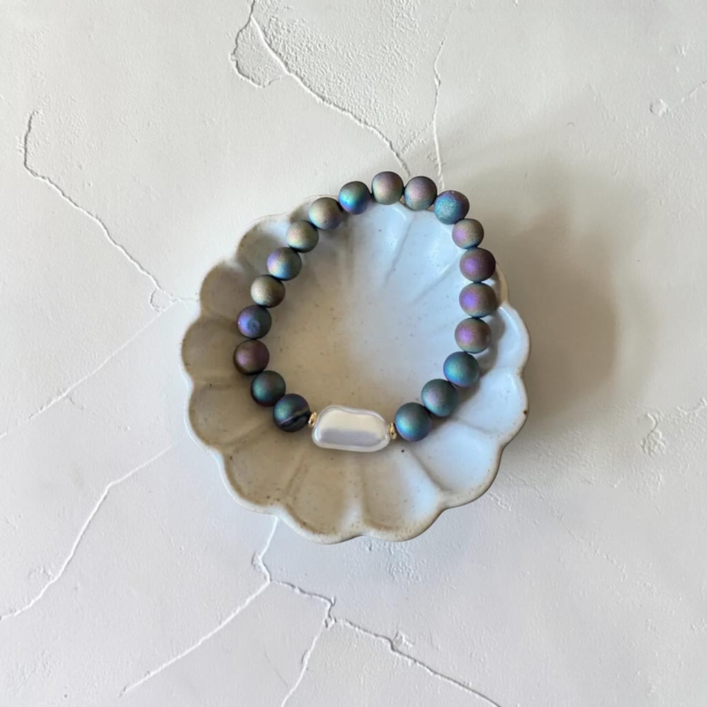 A beaded bracelet with iridescent blue, purple, and gray beads, featuring a central white stone accent, displayed on a scalloped ceramic dish against a textured off-white background.