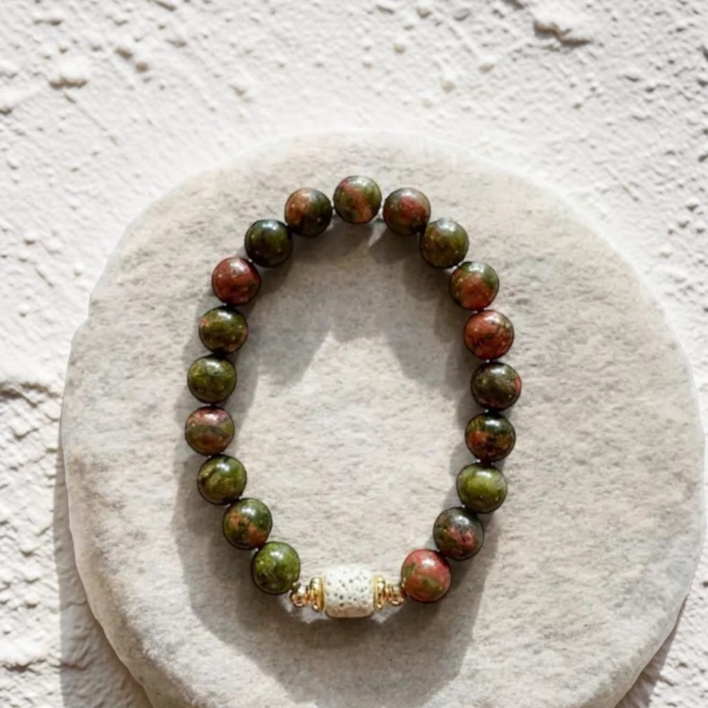 A beaded stretch bracelet made of round green and reddish-brown stone beads, with a white textured spacer bead and small gold-toned accents, displayed on a round light beige stone slab against a textured off-white wall.
