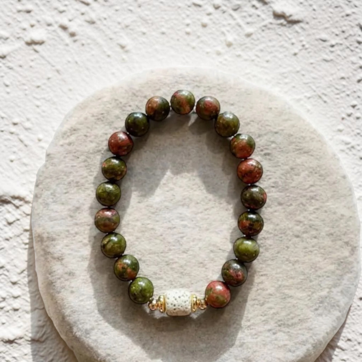 A beaded stretch bracelet made of round green and reddish-brown stone beads, with a white textured spacer bead and small gold-toned accents, displayed on a round light beige stone slab against a textured off-white wall.