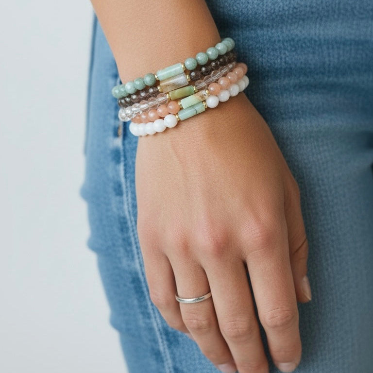 A close-up of a person's hand wearing a stack of five beaded stretch bracelets in pastel colors, paired with a silver band ring and blue jeans.