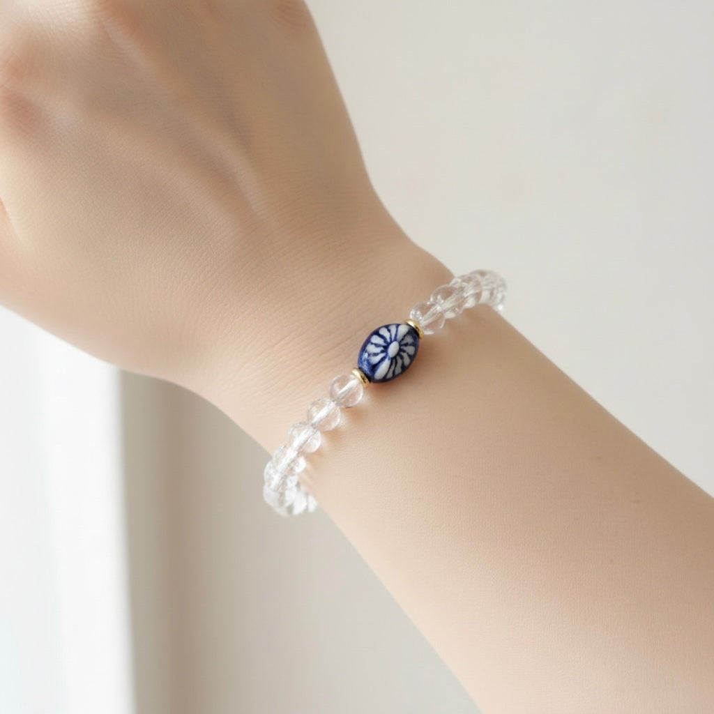 A clear faceted beaded bracelet worn on a wrist, featuring a central round blue and white floral or sunburst bead.