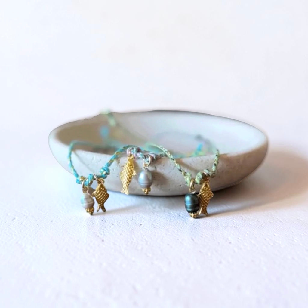 string bracelet pieces with small gold-toned charms and pale beads, draped over a small round ceramic dish on a white surface.