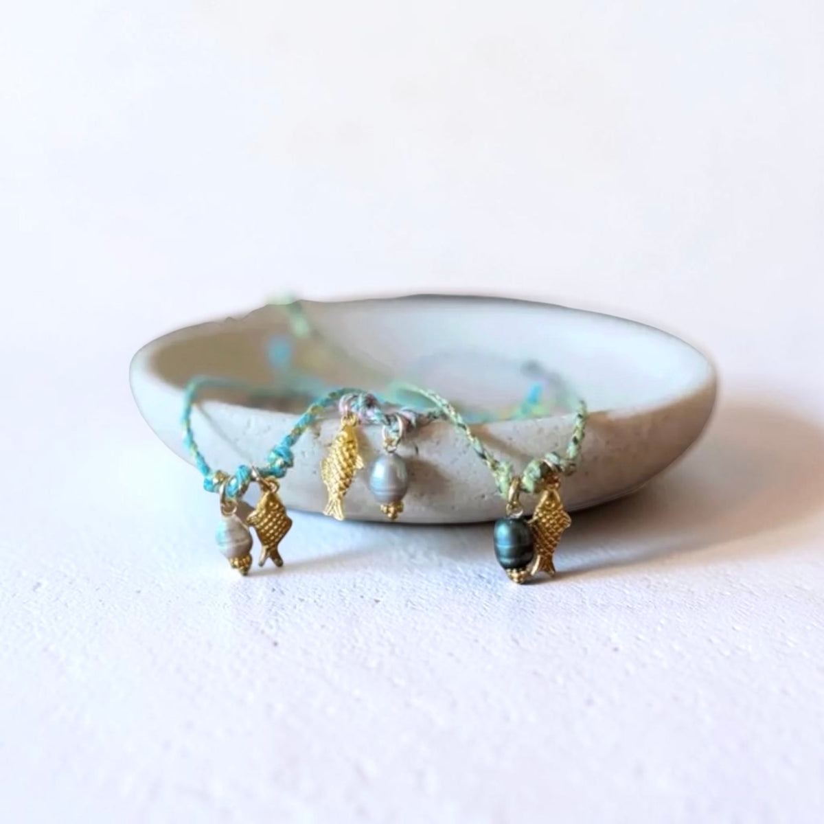 string bracelet pieces with small gold-toned charms and pale beads, draped over a small round ceramic dish on a white surface.