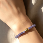 A close-up of a person's wrist wearing a beaded bracelet with lavender-purple round beads and a central faceted gold accent bead, set against a soft neutral fabric background.