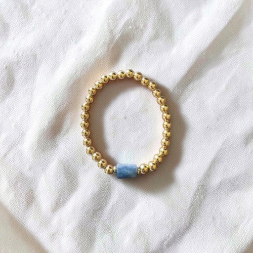 A beaded stretch bracelet with small gold-toned beads and a single light blue rectangular accent bead, photographed on a white fabric background.