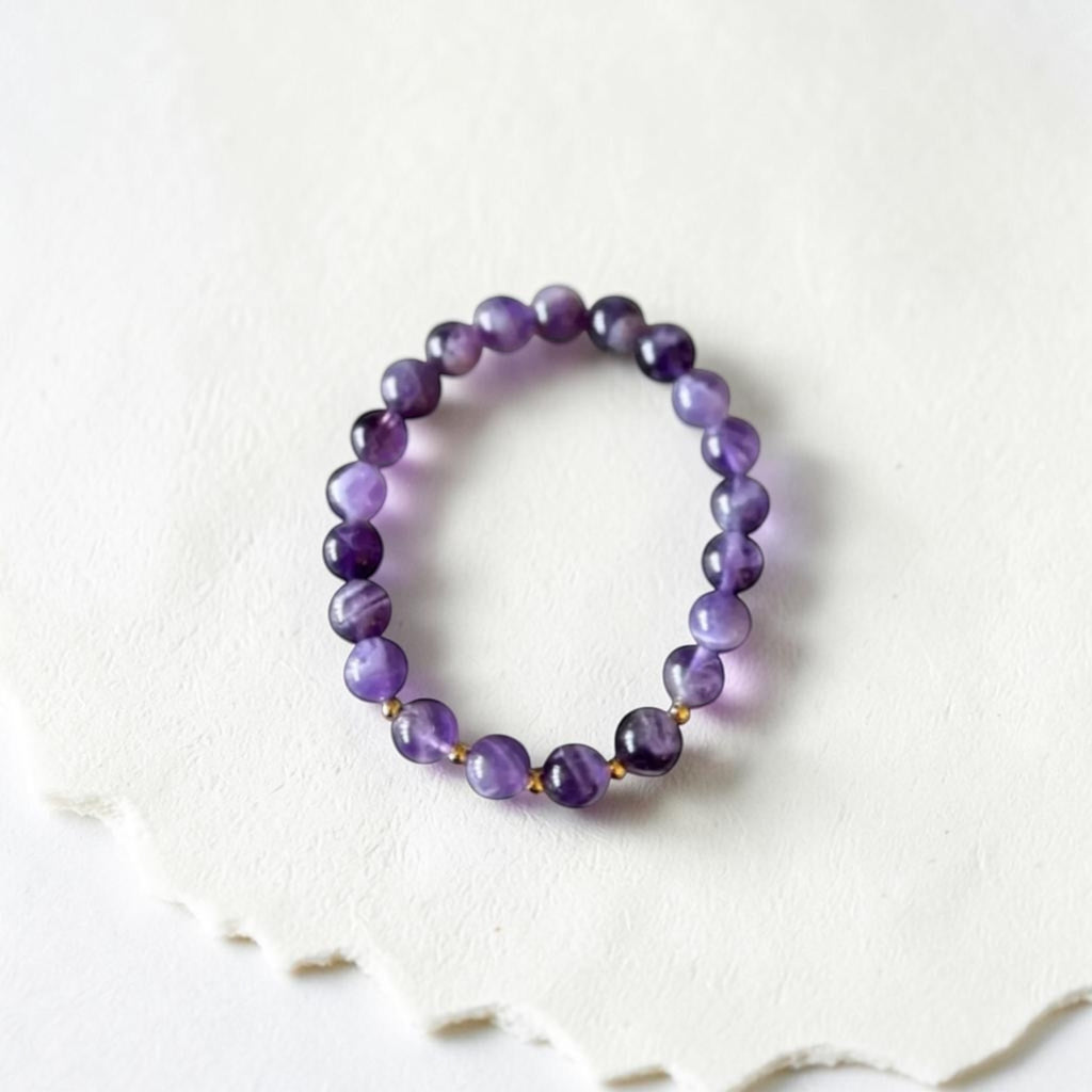A beaded stretch bracelet made of polished purple amethyst and black tourmaline beads separated by small gold spacer beads, displayed on a textured white surface.