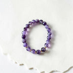A beaded stretch bracelet made of polished purple amethyst and black tourmaline beads separated by small gold spacer beads, displayed on a textured white surface.